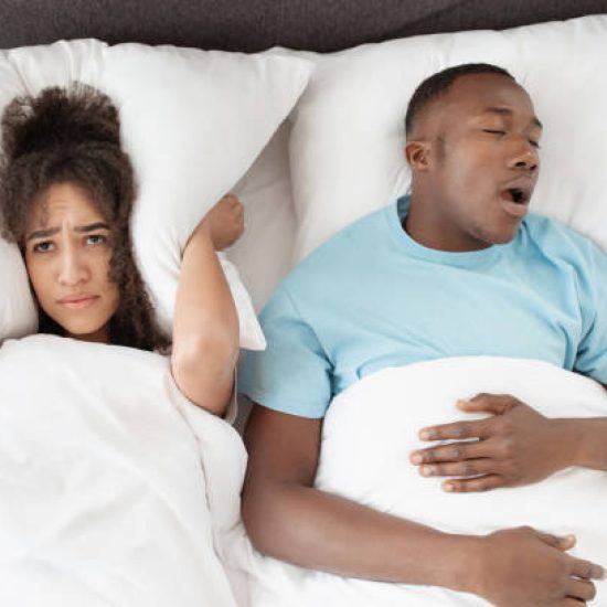 Snoring man sleeping with apnea and sleepless woman, angry wife unable to get to sleep. Upset young african american lady covers ears with pillow so as not to hear noise from snoring guy in bedroom